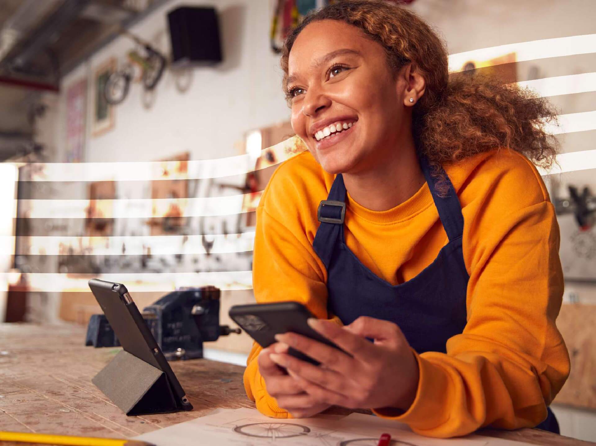 Smiling woman in an orange shirt and blue apron uses a tablet and smartphone at a workshop desk with drawings and tools nearby.
