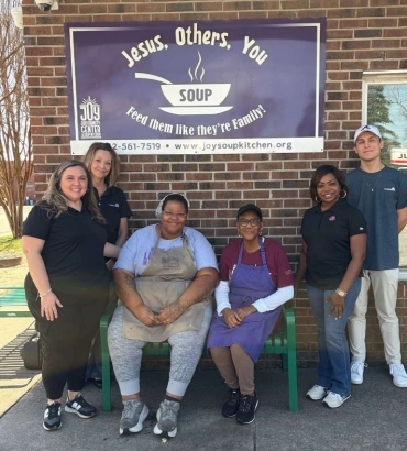 Six volunteers pose outside a brick building under a blue Joy Soup Kitchen banner that reads 'Jesus, Others, You' and 'Feed them like they're family.'