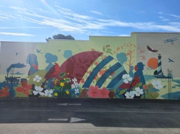Colorful mural on a city wall featuring flowers, birds, and abstract shapes under a bright blue sky with a parking lot in the foreground.