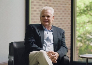 Portrait of an older man with white hair, wearing a dark blazer and light shirt, seated in a black leather chair in a modern office, smiling at the camera.