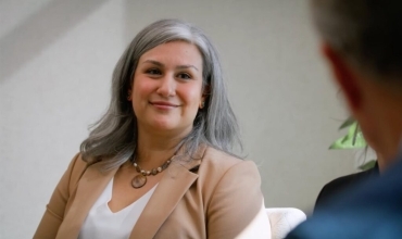 Smiling woman with silver hair wearing a beige blazer, engaged in a conversation with someone off-camera.