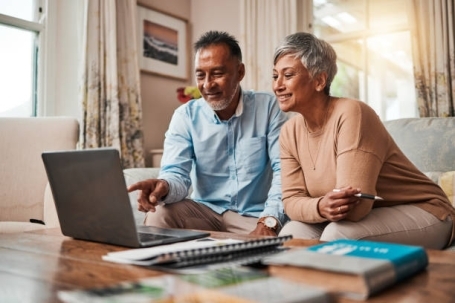Two seniors sit together on a living room sofa, smiling as they look at a laptop screen. They have notebooks on a coffee table in front of them.