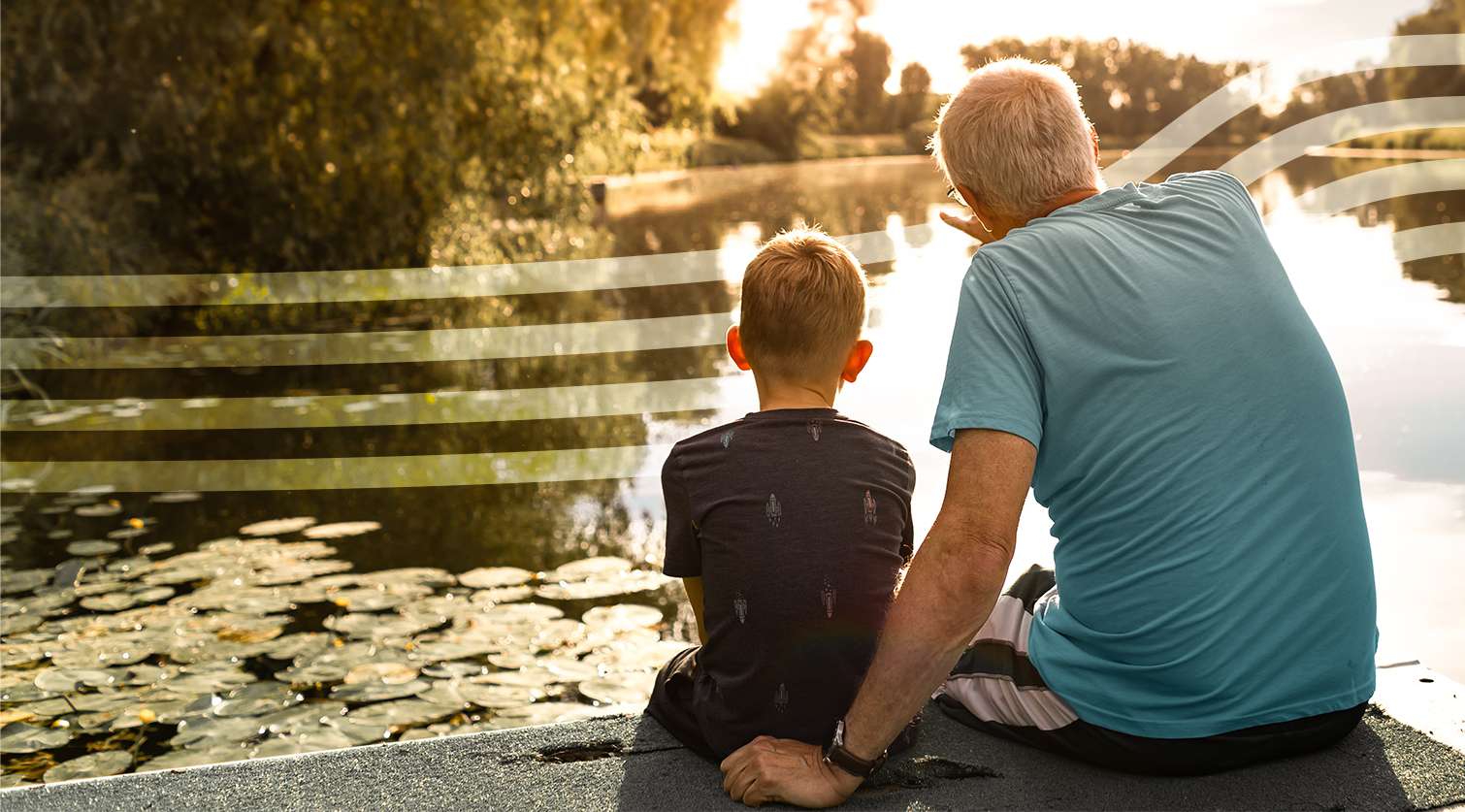 Grandfather and grandson sit on a lakeside edge, watching a sunset over a lily-filled pond.