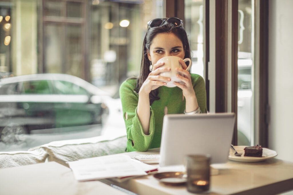 Woman in a green sweater sipping from a large mug at a café table with a laptop nearby and a chocolate cake on the side plate in front of her.