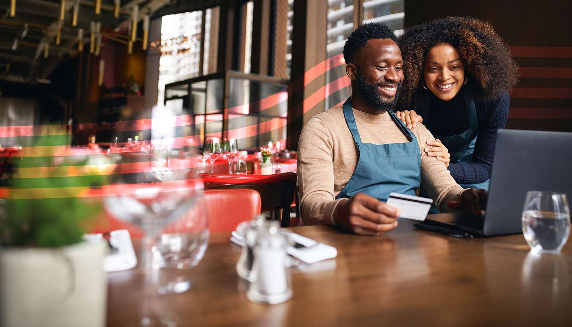 Man in a blue apron sits at a restaurant table using a laptop, holding a card, with a smiling coworker beside him.