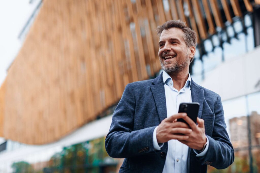 Smiling man in a navy blazer holding a smartphone outdoors against a wood-paneled building exterior.