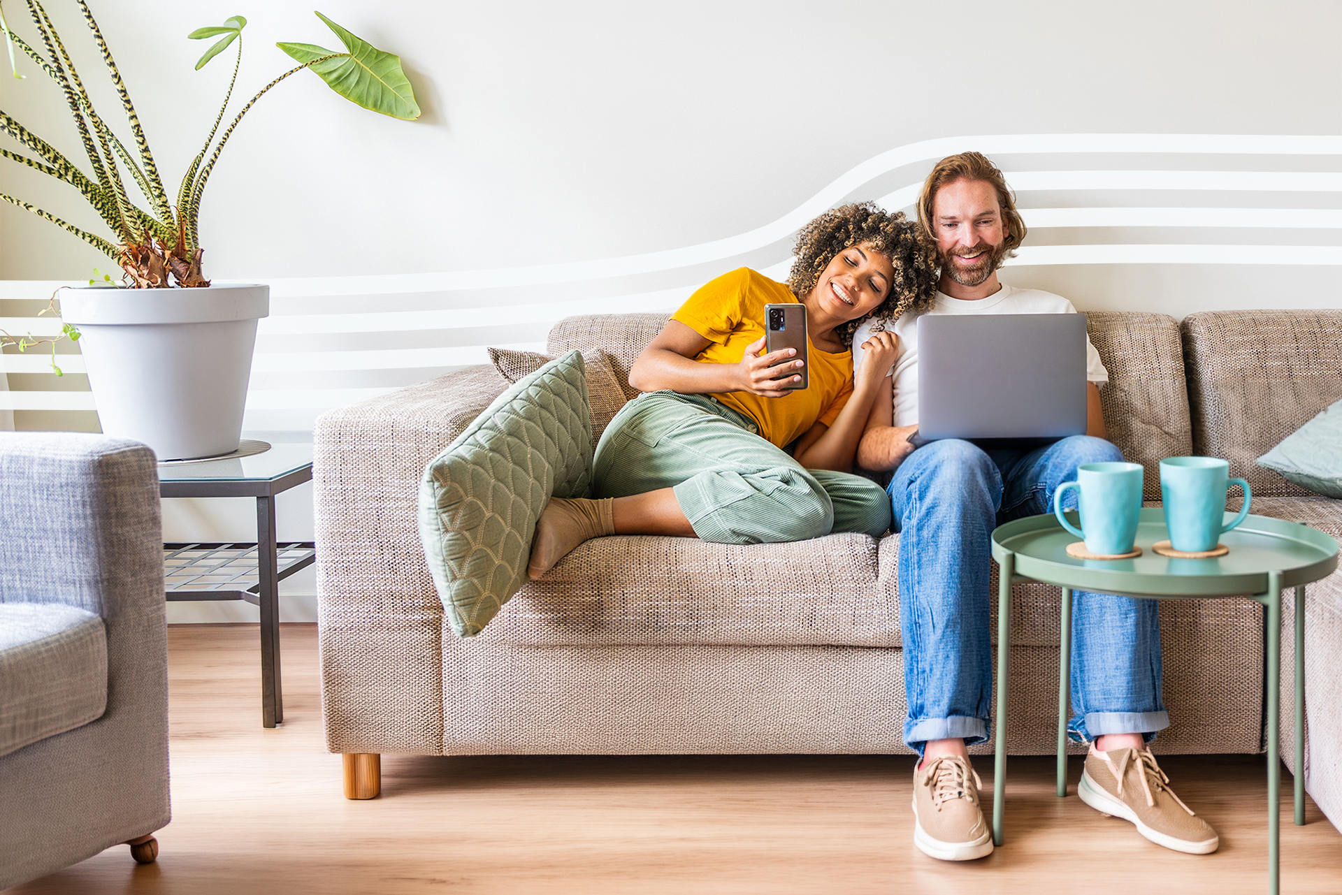 Couple relaxing on a beige sofa in a living room; woman taps a phone, man works on a laptop beside her with a plant nearby.