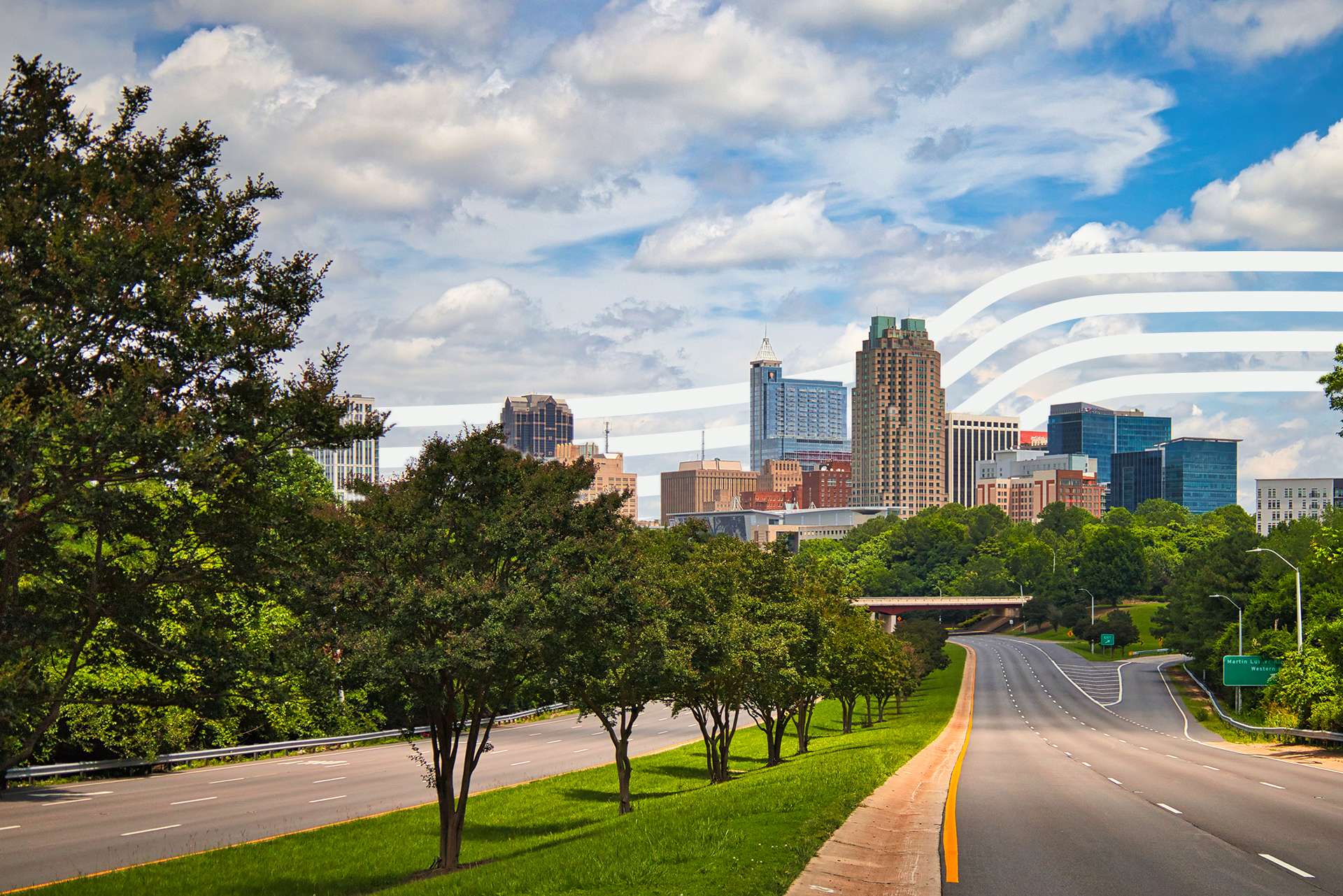 City skyline behind a tree-lined highway under a partly cloudy blue sky, with green foreground trees and grassy median in the foreground.