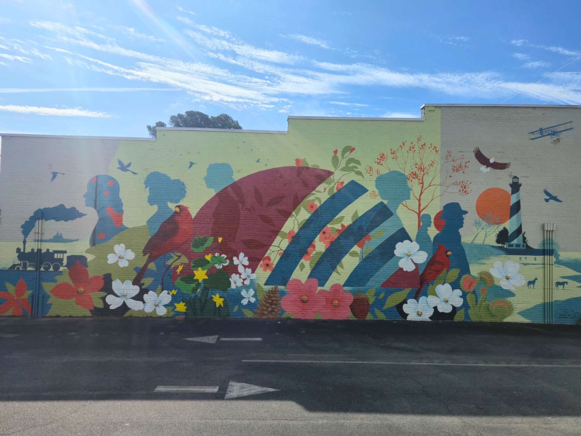 Colorful mural on a city wall featuring flowers, birds, and abstract shapes under a bright blue sky with a parking lot in the foreground.