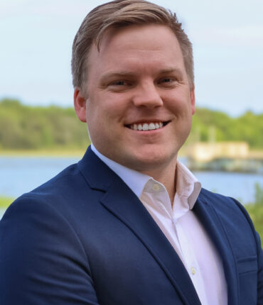Close-up portrait of a smiling man in a navy blazer and light dress shirt, outdoor lake and greenery in the background.