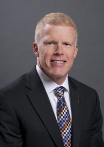 Professional headshot of a smiling man in a dark suit, white shirt, and colorful patterned tie, against a gray studio background.