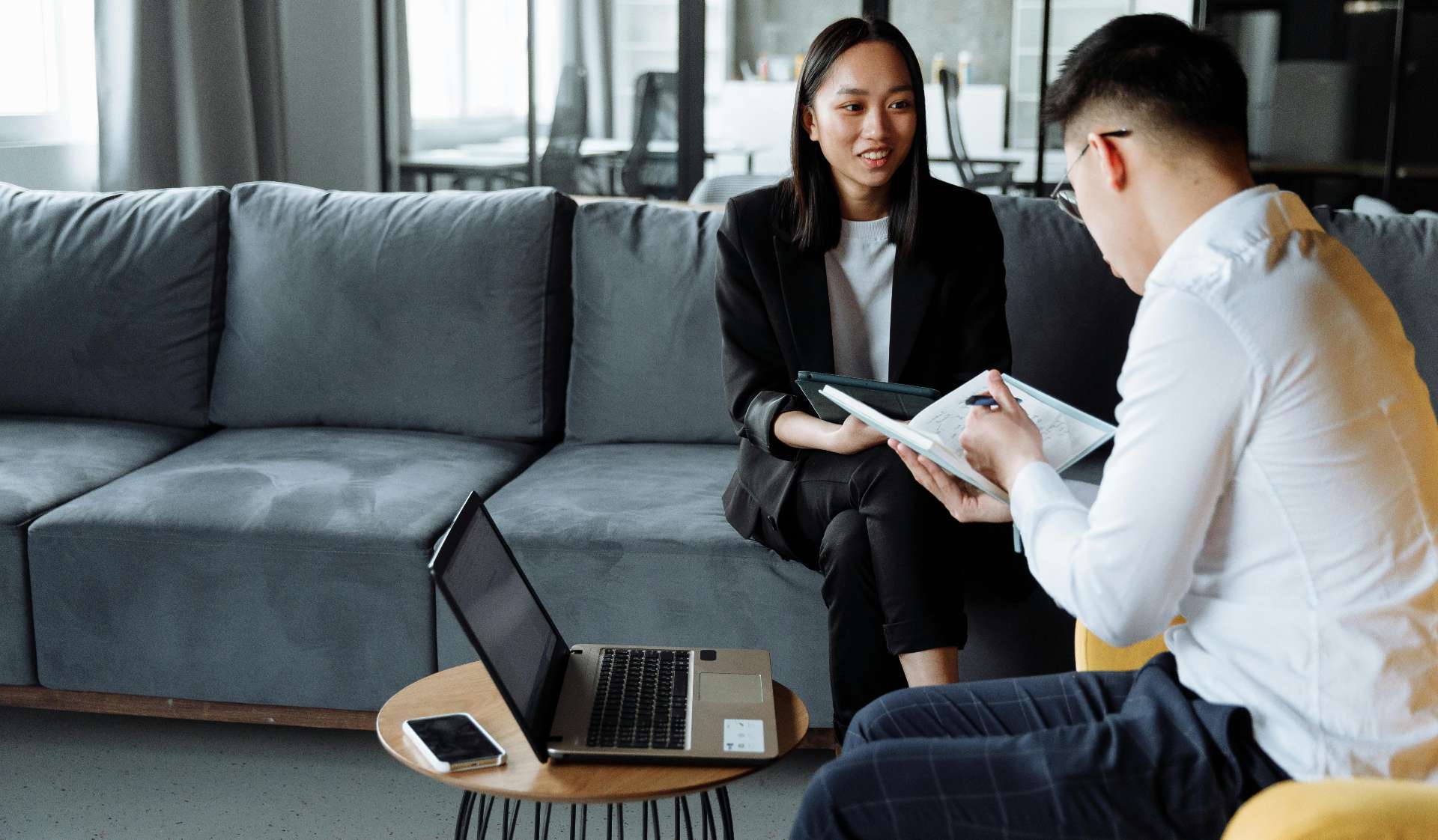 Two coworkers in a modern lounge review documents together, with a laptop and phone on a coffee table.