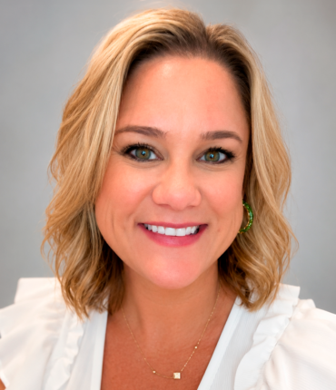 Smiling blonde woman in a white blouse with gold jewelry, close-up portrait against a neutral background.