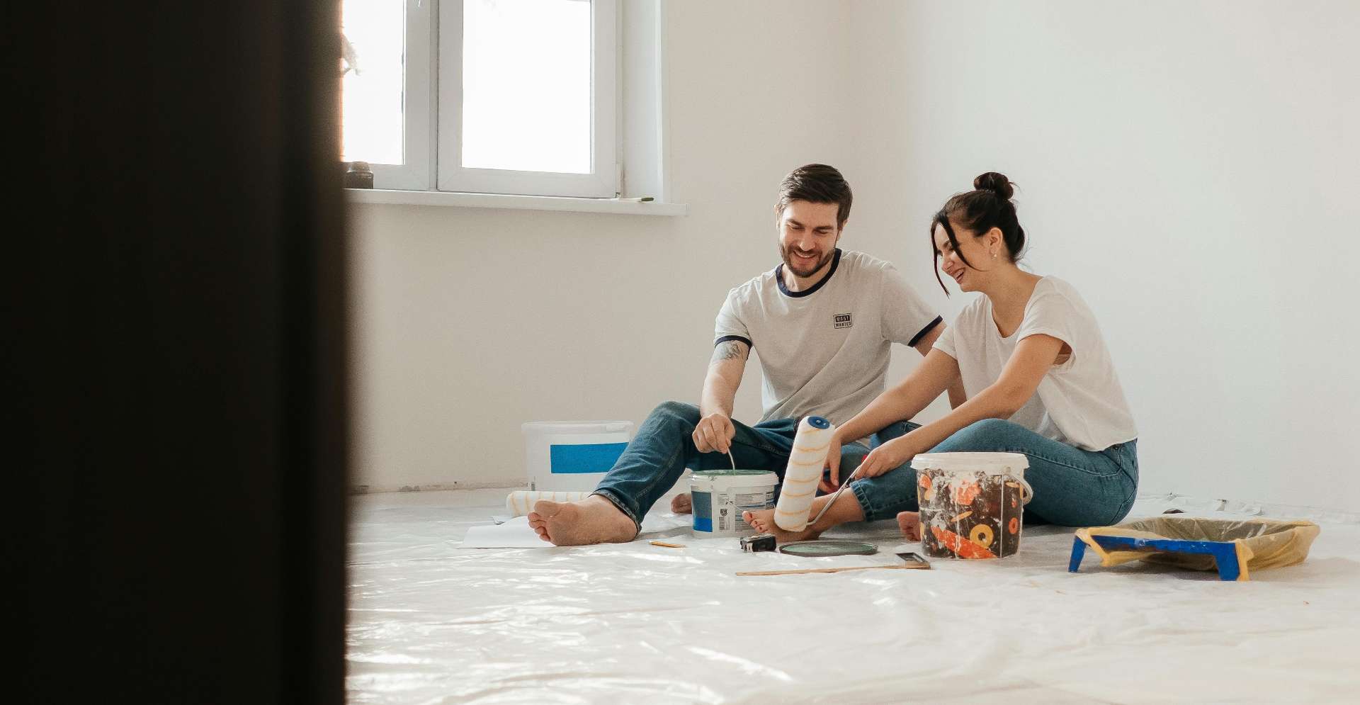 Man and woman sit on a drop-cloth covered floor, painting a room with rollers and paint cans nearby.