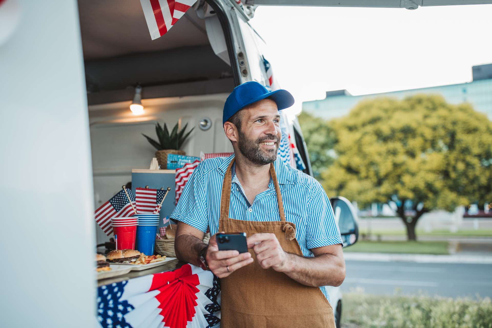 Smiling man in a blue cap and brown apron behind a food truck counter, looking at his phone with red, white, and blue decorations nearby.