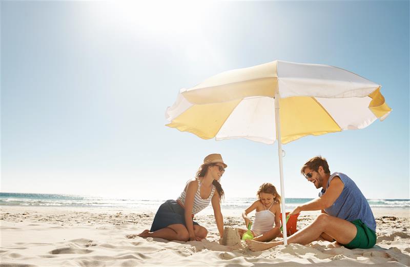 A family of three sits on the sand under a yellow-and-white beach umbrella, playing with sand at the seaside.